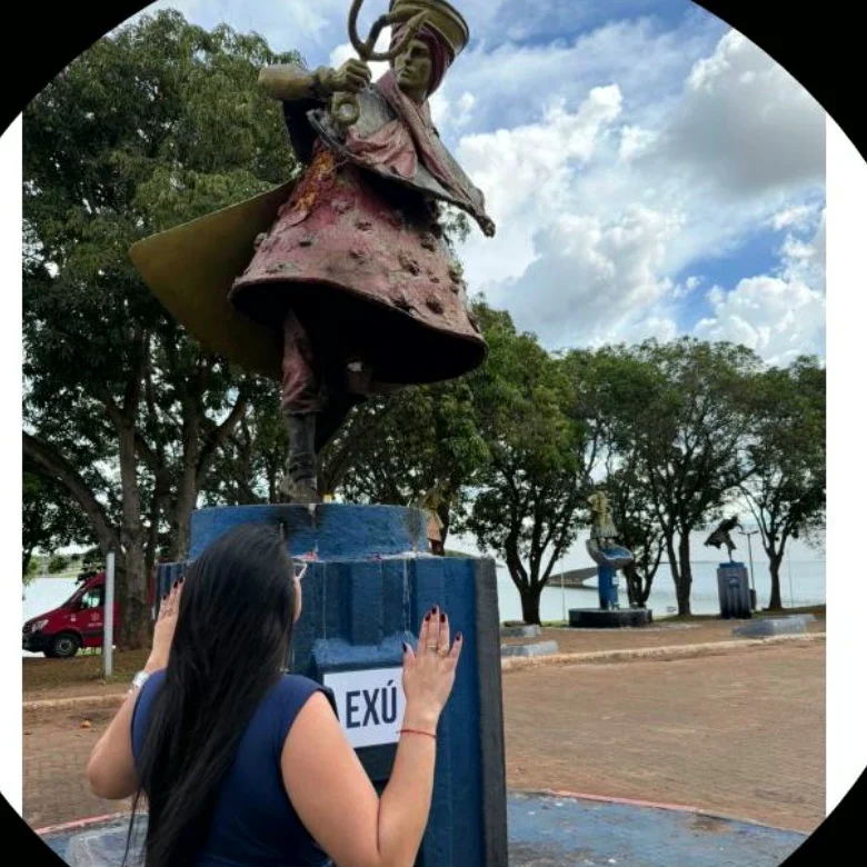 Débora de costas, com as duas mãos apoiadas no pedestal azul de uma estátua de Exú, em um largo à beira do lago.
